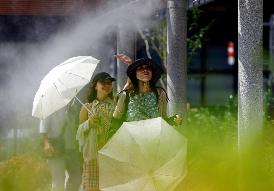 東京で日傘をさす人々がミストシャワーの下で涼んでいる。この日は非常に暑く、日本の環境省は熱中症警戒アラートを発表した。日傘や雨傘の使用で体感温度は下げられると専門家は言う。（PHOTOGRAPH BY ISSEI KATO, REUTERS）