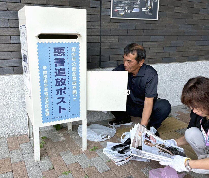 「白ポスト」を開けて中身を回収する前田さん（左）と中西さん。この日は成人向け雑誌に交じって、一般向けの雑誌の切り抜きも多く入っていた（甲賀市、ＪＲ甲南駅前）