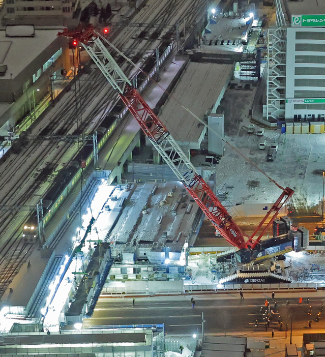 電車が通過する高架の脇で、新幹線駅建設のため大きな橋桁をつり上げる大型クレーン＝9日午後11時50分、札幌市中央区北5西2JRタワーホテル日航札幌から（小松巧撮影）