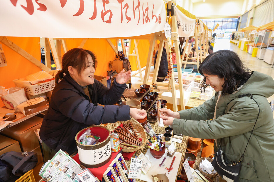 石川県輪島市の食品スーパー「ワイプラザ輪島」に並ぶ輪島朝市の臨時店舗。「漆の店たけはら」の竹原多鶴さん（左）は、「あっという間の一年だった。いっぱい助けてもらえて感謝している」などと振り返った＝２９日