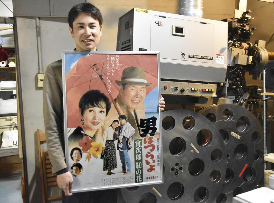 上映に使うフィルム映写機の前で映画「男はつらいよ」のポスターを手にする小山岳志さん＝23日、神戸市兵庫区