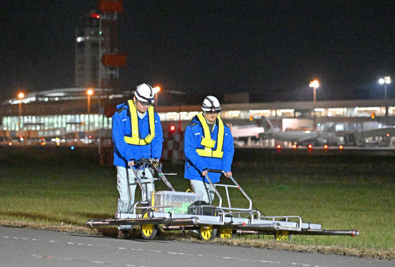 深夜の福岡空港の滑走路周辺で、磁気探査機を使って不発弾の有無を調査する作業員。後方は国際線ターミナル＝福岡市博多区で2024年12月17日午後11時53分、野田武撮影