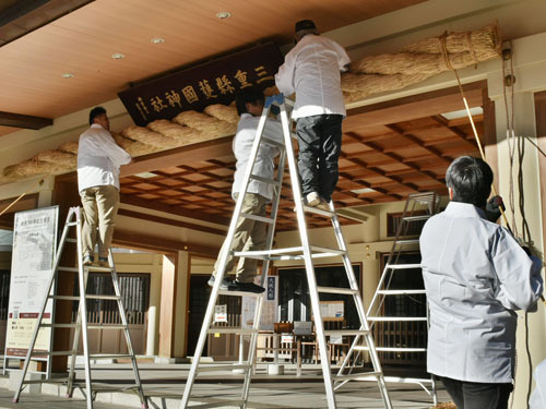 【大しめ縄を縄で引き上げ取り付ける会員ら＝津市広明町の県護国神社で】