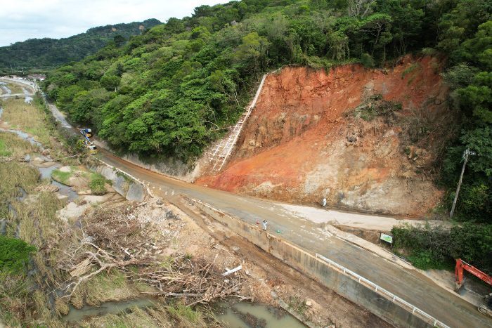 氾濫した比地川（手前）と土砂崩れ現場。路上の土砂の撤去は進んだ。左奥に比地区の住宅地が見える＝６日、国頭村比地（小型無人機で小川昌宏撮影）