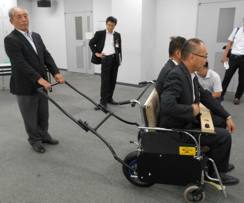 [写真]避難者2人を運べる「車輪付きベンチ」と、引き手を持つ米澤代表（長野県庁で）